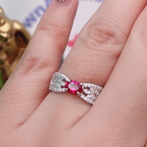 May include: A silver ring featuring a red gemstone surrounded by smaller white gemstones. The ring is worn on a person's finger.