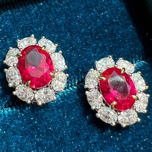 Puede incluir: Un par de pendientes de rubí y diamantes en una caja de joyería de terciopelo turquesa. Los pendientes presentan un gran rubí ovalado rodeado por un halo de diamantes más pequeños.