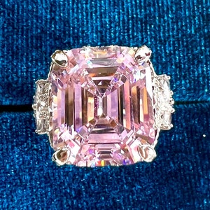 May include: A pink square-shaped engagement ring with a large, clear, pink gemstone surrounded by smaller, clear gemstones. The ring is sitting in a blue velvet ring box.