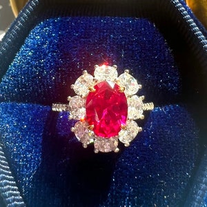 Op de afbeelding: Een ring met een rode edelsteen met een halo van kleinere witte edelstenen, gezet in een gouden band. De ring wordt gepresenteerd in een blauwe fluwelen ringdoos.