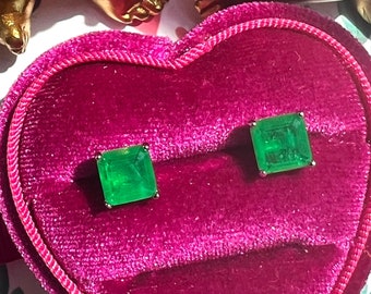 Puede incluir: Un par de pendientes cuadrados de color verde esmeralda en una caja de terciopelo en forma de corazón. La caja es rosa y tiene un forro de satén blanco.