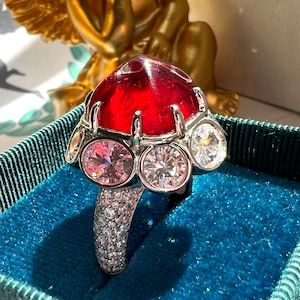 Op de afbeelding: Een zilveren ring met een grote rode edelsteen omringd door kleinere heldere en roze edelstenen. De ring ligt in een blauwe fluwelen doos.