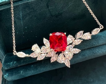 May include: A red gemstone necklace with a large, red, cushion-cut stone surrounded by smaller, clear, marquise-shaped stones. The necklace is on a silver chain and is displayed in a green velvet box.
