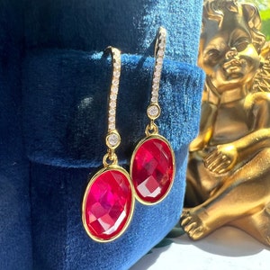 Puede incluir: Un par de pendientes de oro con una piedra ovalada roja y una línea de piedras de circonita cúbica transparentes. Los pendientes están sobre un fondo de terciopelo azul.