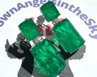 May include: A pair of emerald green statement earrings with a rectangular shape. The earrings have a smaller emerald green stone on top of a larger emerald green stone. Both stones are set in silver.