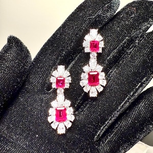 Może przedstawiać: Para kolczyków wiszących z czerwonym kamieniem szlachetnym i gromadą przezroczystych kamieni szlachetnych. Kolczyki leżą na czarnym aksamitnym podłożu.