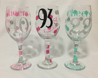 May include: Three wine glasses with silver glitter and pink, teal, and black polka dots. The glasses are decorated with the text "Cheers to 40", "93 & Fabulous", and "Cheers to 29".