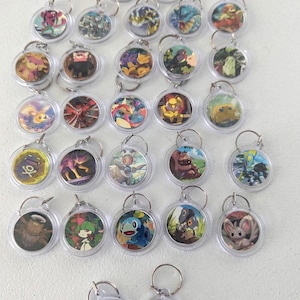 May include: A collection of twenty-seven circular keychains, each featuring a unique colorful character illustration or elemental symbol encased in clear plastic with a metal ring attached to the top for hanging or attachment purposes.