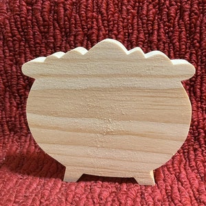 May include: A wooden cauldron shaped craft blank, ready to be painted or decorated. The cauldron is made of unfinished light wood and has a rounded bottom and a wide, flat top.