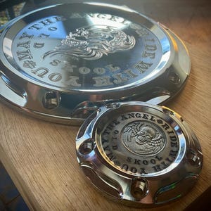 May include: Two polished chrome motorcycle parts with engraved text and a skull design. The larger part is circular, and the smaller part is also circular. The text reads "MAY THE ANGEL RIDE WITH YOU SKOOL".