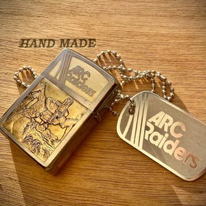 Może przedstawiać: Mosiężny zapalniczka i zestaw nieśmiertelników z napisem "ARC Raiders". Zapalniczka ma grawerowany wzór postaci z bronią. Nieśmiertelnik wisi na łańcuszku. Nad zapalniczką widnieje napis "HAND MADE".