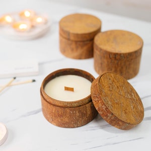 Op de afbeelding: Drie houten kaarsenpotten met deksels. De potten zijn gemaakt van natuurlijk hout met een rustieke afwerking. De kaarsen zijn wit en hebben houten lonten. De tekst "TONKIN HANDICRAFT" is zichtbaar in de linkerbovenhoek van de afbeelding.