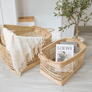 May include: Two woven seagrass storage baskets with handles. The larger basket is filled with a white blanket and the smaller basket contains a magazine with the word "LOEWE" on the cover.
