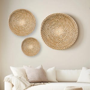 May include: Three round woven wall decorations of varying sizes, crafted from natural materials, hanging on a white wall. The largest piece is in the centre, with a smaller piece to the left and the smallest piece to the left of the centre piece. The wall decor is in a living room setting with a white sofa and a wooden coffee table.