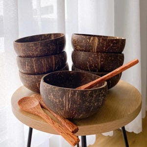 Peut inclure: Un ensemble de six bols en noix de coco brun avec un motif de grain de bois naturel, empilés les uns sur les autres. Deux cuillères en bois sont placées sur une table en bois à côté des bols.