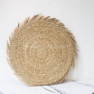 Puede incluir: Un mantel individual redondo de paja trenzada con un borde de flecos. El mantel individual está hecho de materiales naturales y tiene un color marrón claro.