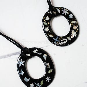 May include: Two black oval pendants with floral and bird designs. The pendants are adorned with white and gold accents, and are suspended from black cords. The pendants are on a white marble surface. The text "TONKIN HANDICRAFT" is visible.