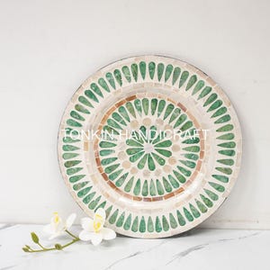 May include: A round decorative plate with a green and white mosaic design. The plate has a circular pattern of green teardrop shapes around a central circle of white and brown mosaic tiles. The text "FONKIN HANDICRAFT" is visible on the plate.