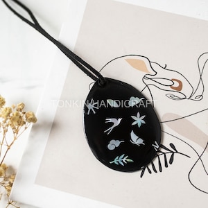 May include: Black oval pendant with a black cord. The pendant features a design of a bird, butterfly, clouds, and leaves. The text "TONKINHANDICRAFT" is printed on the pendant.