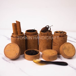 May include: Five small wooden containers with lids, filled with various spices and herbs. The containers are arranged in a row on a white marble surface. The text "TONKIN HANDICRAFT" is visible on the containers.