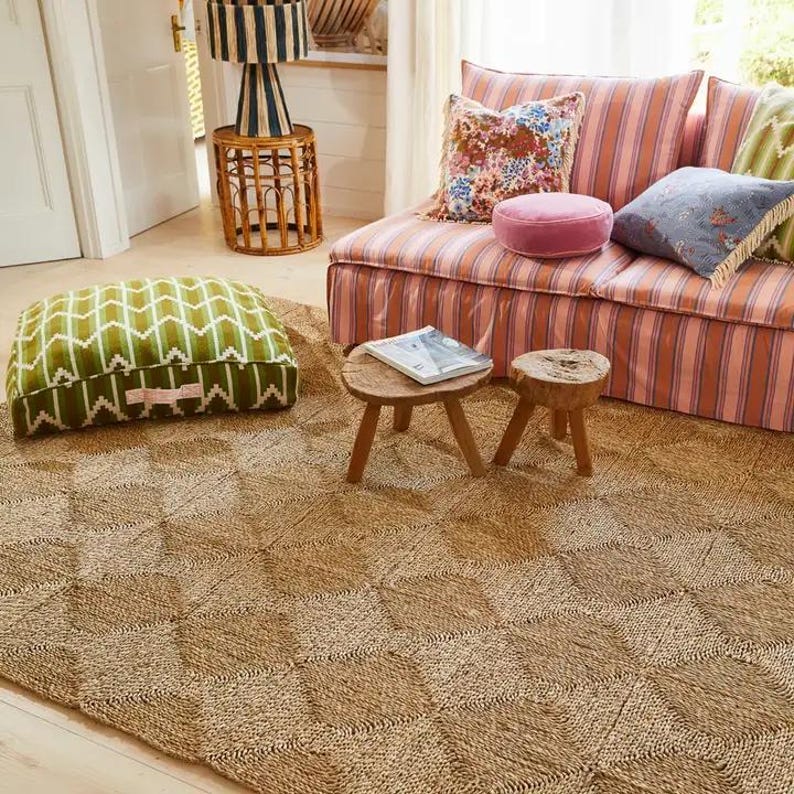 May include: A natural fiber woven rug with a diamond pattern in a light brown color. The rug is in a living room setting with a pink striped sofa, green and white patterned throw pillows, and two wooden stools.