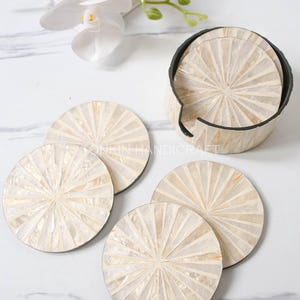 Op de afbeelding: Set van zes witte parelmoer-onderzetters met een zonnestralenontwerp. De onderzetters zitten in een ronde houder met een zwarte rand.
