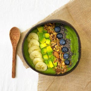 Puede incluir: Un tazón de batido verde con rodajas de plátano, aguacate, granola, arándanos y semillas de chía en un tazón de coco marrón oscuro. Una cuchara de madera está a la izquierda del tazón.