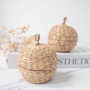 Puede incluir: Dos recipientes de almacenamiento esféricos tejidos con tapas. Los recipientes están hechos de una fibra natural marrón claro y tienen una superficie texturizada. El texto "TONKIN HANDICRAFT" está en la parte superior.