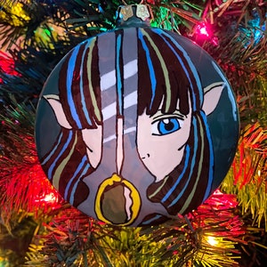 May include: A hand-painted Christmas ornament with a teal background. The ornament features a stylized face with blue eyes, brown and blue hair, and a gold pendant. The ornament is hanging on a Christmas tree with colorful lights.