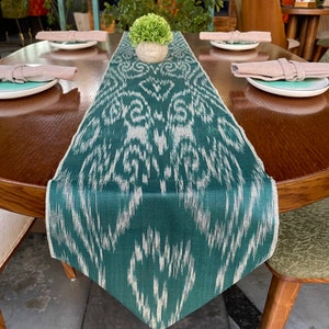 Op de afbeelding: Een groene en witte tafelrunner met een witte en groene plant in een witte pot op een houten tafel.