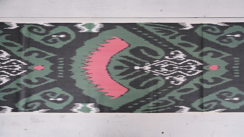 May include: A close-up of a green and black fabric with a pink and white pattern. The fabric has a traditional ikat design.