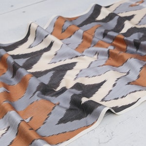Puede incluir: Una tela con un patrón de chevrón repetido. El diseño presenta bandas alternas de gris, negro, marrón y blanco roto. La tela está drapeada, mostrando su textura y la interacción de los colores.