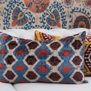 Op de afbeelding: Drie decoratieve kussens met een geometrisch patroon in tinten blauw, rood, geel en wit. De kussens liggen op een witte bank met een bloemmotief op de achtergrond.