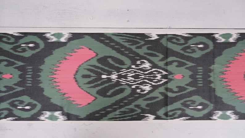 May include: A green, black, and pink patterned fabric with a traditional ikat design. The fabric has a white background with a repeating pattern of swirls and diamonds.