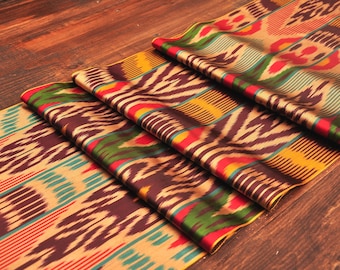 May include: Folded fabric with a vibrant ikat pattern in shades of brown, tan, red, green, and teal. The fabric is arranged on a dark wooden surface, showcasing its intricate design and texture.