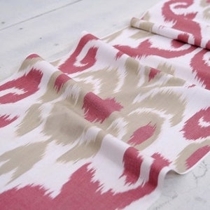 May include: White fabric with a pink and beige ikat pattern. The fabric is folded and draped on a white surface.