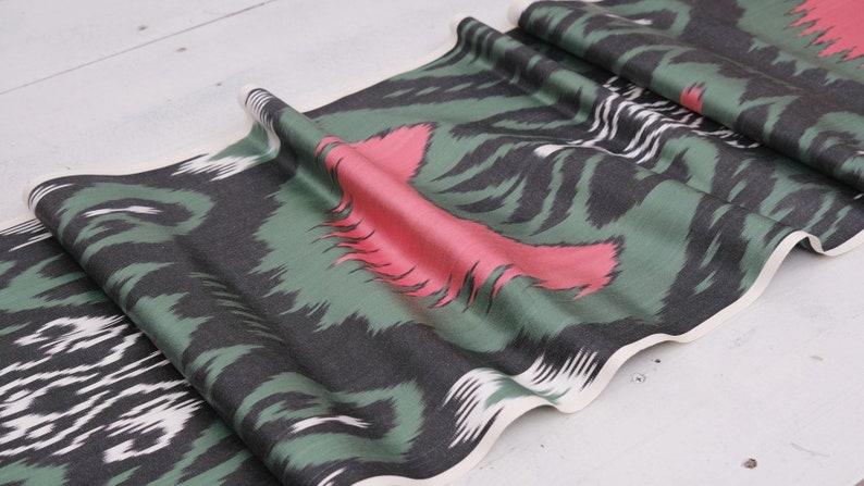 May include: Green, black, and pink patterned fabric with a white border. The fabric is folded and has a wavy edge.
