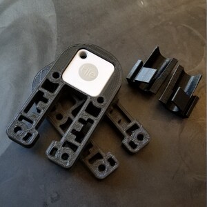 May include: A 3D printed black plastic holder for a Tile Bluetooth tracker. The holder has a cutout for the Tile and a clip to attach it to a belt or bag. The Tile is white with the word "Tile" printed on it.