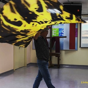 May include: A person wearing a black shirt and blue jeans is holding a large yellow and black fabric flag. The flag is being waved in the air. The person is standing in a room with a projector screen displaying the text "Lord God of Abraham by Paul Wilbur".