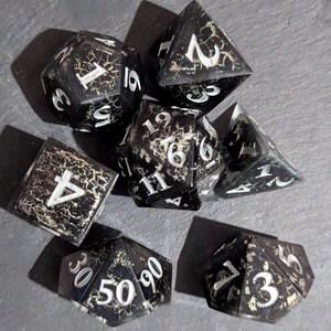 May include: A set of seven black dice with silver numbers and a speckled, aged appearance. The dice are in various shapes and sizes, including a d4, d6, d8, d10, d12, and d20.