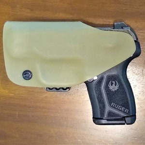 Pu&ograve; includere: Una pistola Ruger nera con un'impugnatura testurizzata e una fondina nera. La fondina &egrave; realizzata in un materiale verde chiaro e ha un clip sul fondo.
