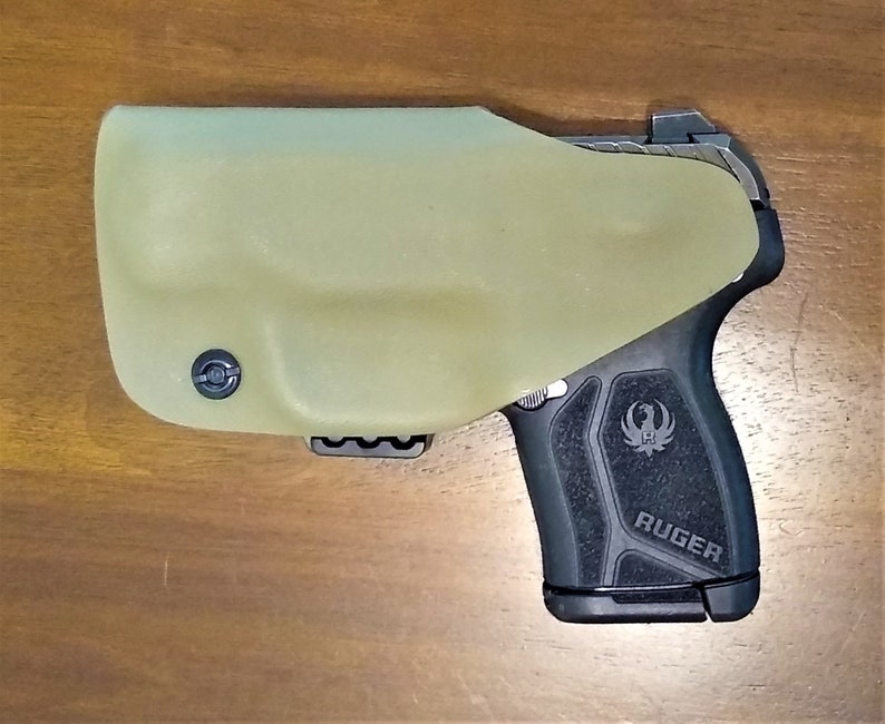 Pu&ograve; includere: Una pistola Ruger nera con un'impugnatura testurizzata e una fondina nera. La fondina &egrave; realizzata in un materiale verde chiaro e ha un clip sul fondo.