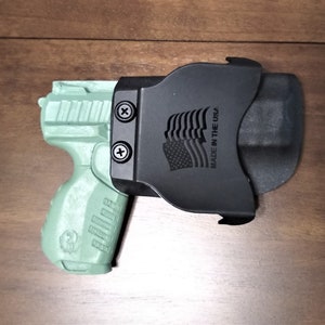 May include: A green handgun with a black holster. The holster has the text "Made in the USA" and an American flag design.