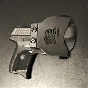 Puede incluir: Una pistola negra con una corredera plateada y una funda de pol&iacute;mero negra. La funda tiene el texto "MADE IN THE USA" y un dise&ntilde;o de bandera estadounidense estilizado.