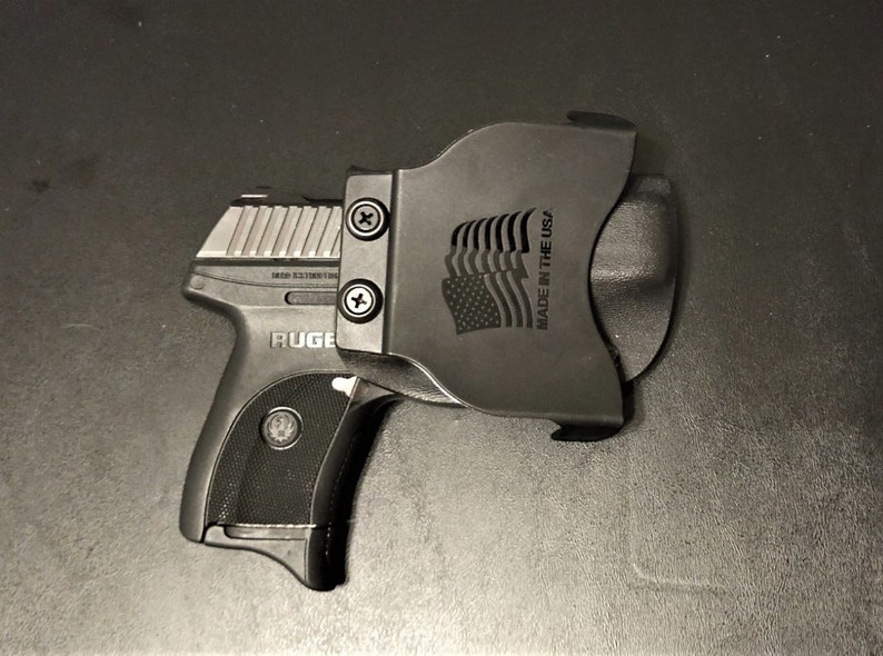 Puede incluir: Una pistola negra con una corredera plateada y una funda de pol&iacute;mero negra. La funda tiene el texto "MADE IN THE USA" y un dise&ntilde;o de bandera estadounidense estilizado.