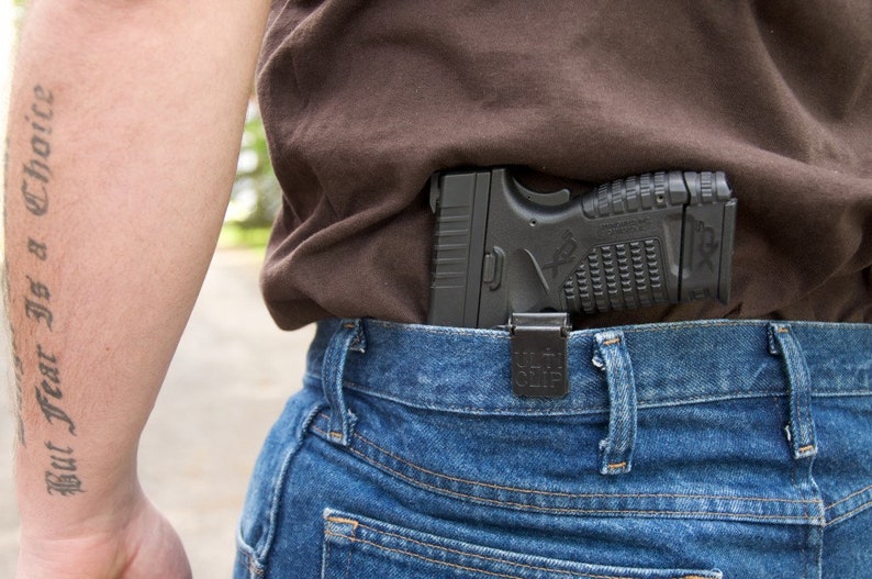 May include: A black handgun is tucked into the waistband of a pair of blue jeans. The gun is secured with a black holster. The person wearing the jeans has a tattoo on their arm that reads "But Fear Is a Choice."