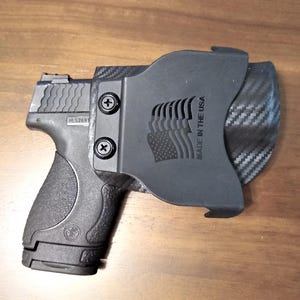 May include: A black polymer gun holster with a carbon fiber pattern and the text "Made in the USA". The holster is designed to hold a small black handgun.
