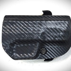 May include: Black carbon fiber holster with a textured weave pattern. The holster features a curved design, a black clip, and a screw. The item is designed to hold a firearm.