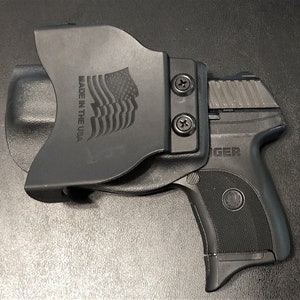 Puede incluir: Una funda de pol&iacute;mero negro con una pistola negra. La funda tiene un clip para cintur&oacute;n y un logotipo de la bandera de Estados Unidos con el texto "Made in the USA". La pistola tiene un armaz&oacute;n de pol&iacute;mero negro y una corredera plateada. La corredera tiene la palabra "GER" en ella.