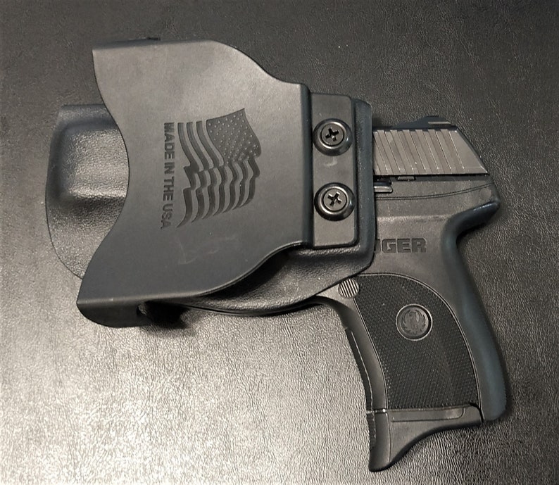 Puede incluir: Una funda de pol&iacute;mero negro con una pistola negra. La funda tiene un clip para cintur&oacute;n y un logotipo de la bandera de Estados Unidos con el texto "Made in the USA". La pistola tiene un armaz&oacute;n de pol&iacute;mero negro y una corredera plateada. La corredera tiene la palabra "GER" en ella.